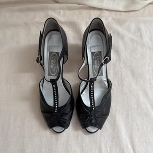 Könnte beinhalten: Ein Paar schwarze Leder-High Heels mit offenem Zehenbereich und T-Steg-Design. Die Absätze haben eine silberne Innensohle und eine Reihe von Strasssteinen. Die Schuhe haben einen verstellbaren Knöchelriemen.