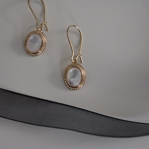 May include: A pair of gold-toned drop earrings featuring oval-shaped, iridescent white cabochons set within a textured, beaded metal border. The earrings are suspended from curved gold-toned ear wires, showcasing a classic and refined design.
