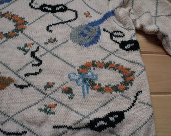 Könnte beinhalten: Cremefarbener Pullover mit Rautenmuster und festlichen Stickereien. Zu den Merkmalen gehören orangefarbene Blumenkränze, schwarze Masken und blaue Schleifen. Der Pullover hat lange Ärmel und einen klassischen Rundhalsausschnitt.
