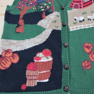 May include: A green, black, and brown knitted vest with a button-up front. The vest features a variety of embroidered designs, including apples, trees, a barn, and sheep.