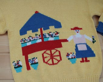 Könnte beinhalten: Ein gelber Strickpullover mit einem blauen und roten Blumenwagen, einer Frau in einem blauen Kleid und einem roten Hut und einem Etikett mit der Aufschrift "Made in Nepal".