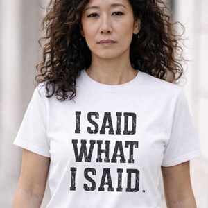 May include: White t-shirt with the text "I SAID WHAT I SAID" in a bold, black, distressed font. The shirt is worn by a person in blue jeans, walking on a paved walkway. The background is blurred, suggesting an outdoor setting.