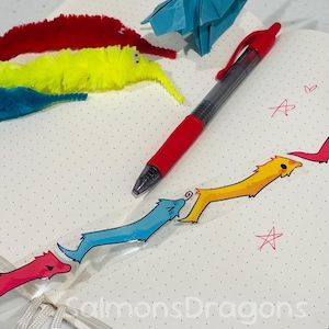 May include: A red pen rests on a dotted notebook next to a line of colorful worm-like illustrations. Three fuzzy worm toys in red, yellow, and blue are also present, along with a blue origami crane.