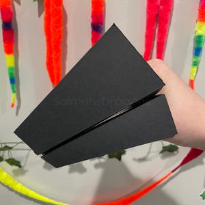 May include: A black, triangular-shaped box with a matte finish. The box is composed of two separate, angled pieces that fit together to form a wedge shape. The text "SalmonsDragons" is printed in a subtle, dark font on the side of the box.