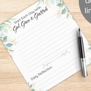 May include: A white notepad with lined pages and a floral design in shades of blue and gold. The notepad has the text "Start Each Day With God, Grace & Gratitude" and "Daily Reflection:" printed on it. A black and silver pen rests on the notepad.