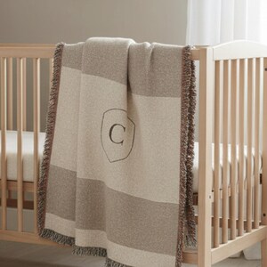 May include: A light wood cot with a cream mattress and a beige and tan striped blanket draped over the side. The blanket features a shield design with the letter "C". Two shelves with stuffed toys are mounted on the wall.