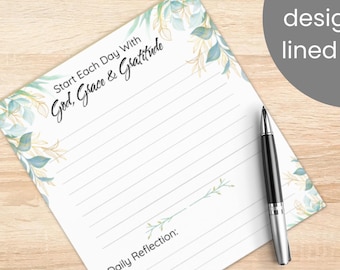 May include: A square notepad featuring a white background with watercolor-style blue and green leaves along the edges. The text reads Start Each Day With God, Grace & Gratitude and Daily Reflection. A black and silver ballpoint pen rests on the paper.