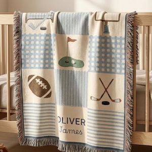 Puede incluir: Una manta tejida con un patrón de temática deportiva que incluye un balón de fútbol americano, un green de golf, palos de hockey y postes de portería. El diseño presenta secciones de cuadros y rayas azul claro con el texto OLIVER James en azul.