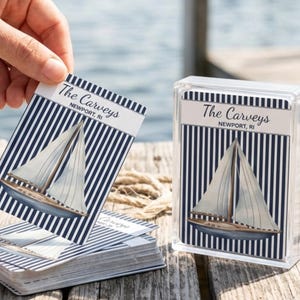 May include: A deck of playing cards featuring a navy blue and white vertical striped pattern with a sailboat illustration and the text The Carveys Newport, RI. The cards are stored in a clear plastic case, with one card held up to display.