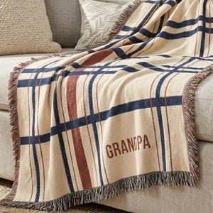 May include: A woven throw blanket featuring a cream-coloured base with a plaid pattern in navy blue and dark red stripes. The word "GRANDPA" is embroidered in dark brown lettering on the lower section. The blanket has fringed edges on the sides.