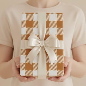 May include: A rectangular gift box wrapped in white and tan gingham patterned paper. The box is tied with a smooth, solid cream-colored satin ribbon that forms a decorative bow centered on the front, creating a clean and classic presentation.