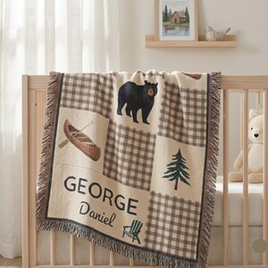 Può includere: Una coperta tessuta con un motivo a quadri color crema e marrone, con pannelli che mostrano un orso nero, una canoa marrone, un pino verde e una sedia Adirondack verde. Il testo GEORGE Daniel è stampato in nero sulla coperta.