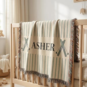 Può includere: Coperta per bambini color crema con strisce verticali verde chiaro e il nome "ASHER" in lettere nere. La coperta presenta un paio di sci su ogni lato del nome. La coperta ha un bordo sfrangiato ed è drappeggiata su una culla di legno.