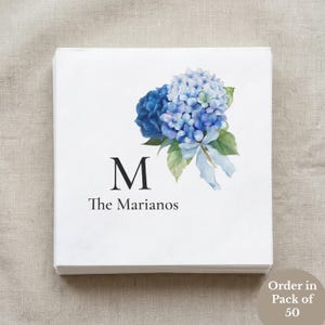 May include: A square white paper napkin featuring a watercolor illustration of blue and light blue hydrangea flowers with green leaves and a light blue ribbon. The letter M is printed in black, followed by the text The Marianos below it.