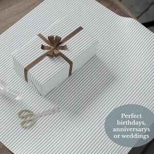 May include: A wrapped gift box with a brown ribbon and bow sits on striped wrapping paper. The wrapping paper has a blue and white striped pattern. A pair of gold-handled scissors is next to the gift.