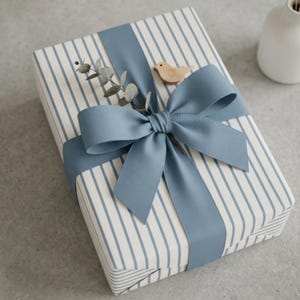 May include: A wrapped gift with blue and white striped paper, tied with a wide blue ribbon and bow. A small wooden bird and sprig of greenery are tucked under the bow, creating a decorative and elegant presentation.