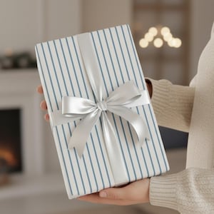 May include: A wrapped gift with blue vertical stripes on white paper, tied with a silver satin ribbon and bow. The gift is held in front of a blurred background.