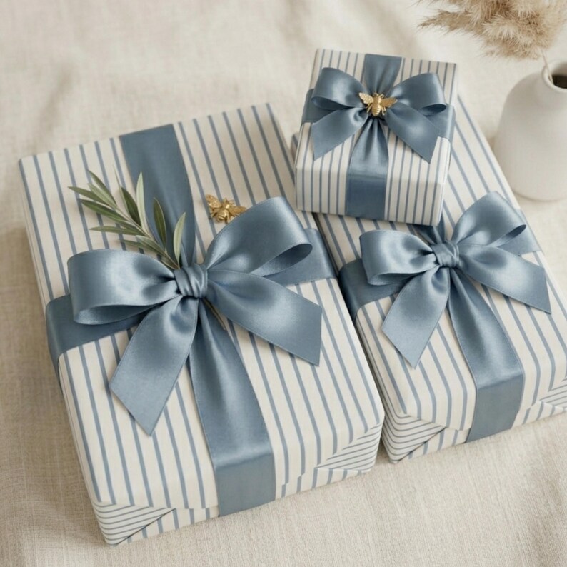 May include: Three wrapped gifts with blue and white striped paper and satin blue bows. One gift has a gold bee embellishment. Another has a sprig of greenery. The gifts are arranged on a white surface.