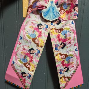 May include: A large hair bow made of white grosgrain ribbon featuring a repeating pattern of various cartoon princesses in colorful dresses. A central cutout of a princess in a blue gown is attached, and the bottom edges are lined with colorful rhinestones.