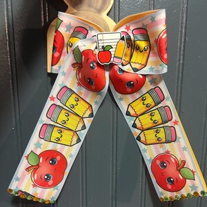 May include: A hair bow made of grosgrain ribbon featuring a pattern of smiling cartoon apples and yellow pencils on a background of pastel stripes and small stars. A small charm depicting a notebook, an apple, and a pencil sits at the center.