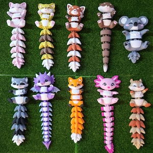 Può includere: Una collezione di giocattoli di animali colorati e articolati, tra cui un gatto rosa, un leopardo giallo e un panda rosso. Ogni giocattolo ha un corpo e una coda segmentati, esposti su una superficie verde.