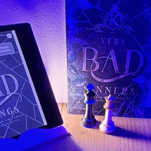 Könnte beinhalten: Ein schwarzer und weißer Schachkönig und eine -königin stehen vor einem Buch mit dem Titel "Very Bad Sinners" und einem E-Reader. Das Buchcover ist dunkelblau mit weißem Text. Der E-Reader zeigt Text aus einem Buch an.