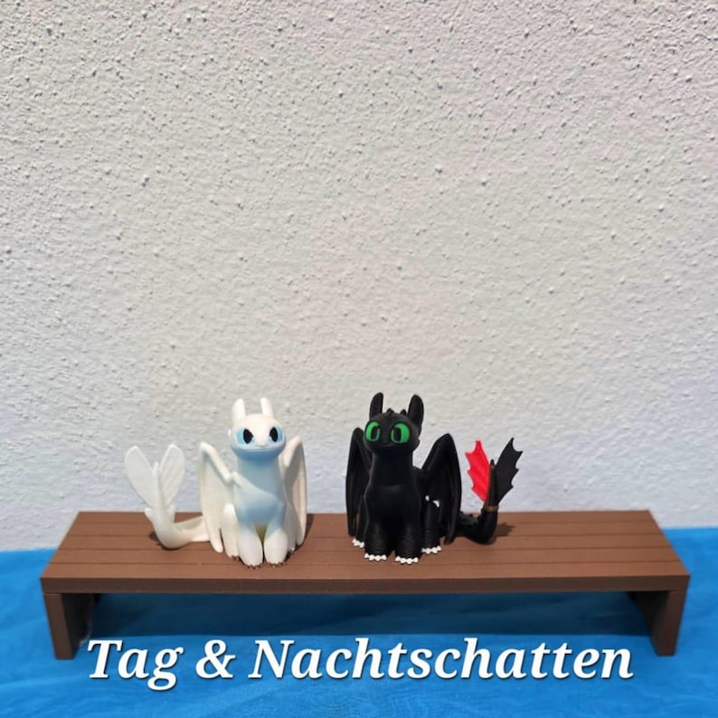 May include: Two dragon figurines, one white and one black, are positioned side by side. The white dragon has blue eyes, while the black dragon features bright green eyes and a single red tail fin. The text Tag & Nachtschatten is displayed below.