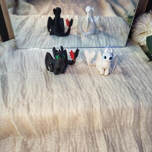 May include: Two small dragon figurines, one black with green eyes and a red tail fin, and one white with blue eyes. The black dragon has small white claws on its feet. Both figures are depicted in a seated position, reflecting in a mirror behind them.