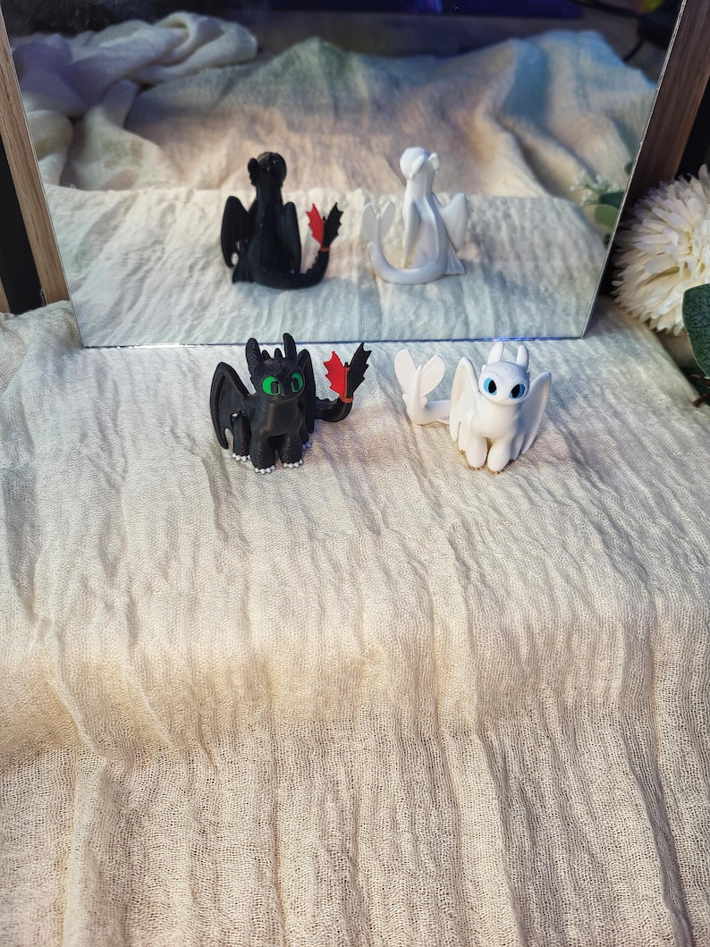 May include: Two small dragon figurines, one black with green eyes and a red tail fin, and one white with blue eyes. The black dragon has small white claws on its feet. Both figures are depicted in a seated position, reflecting in a mirror behind them.