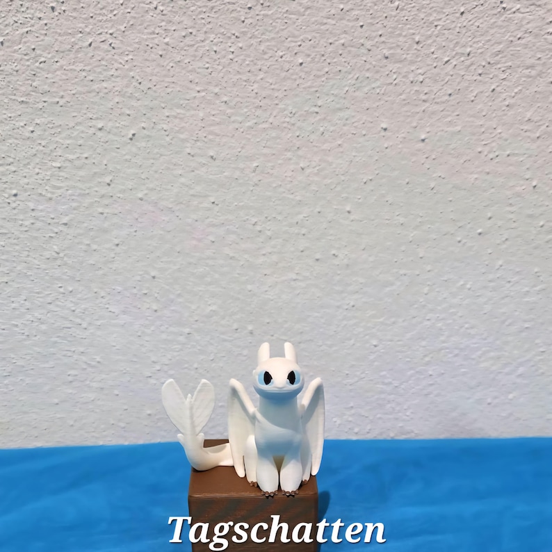 May include: A white dragon figurine with large ears, wings, and a tail, featuring light blue eyes and a blue-tinted snout. The creature is positioned on a brown wooden block. The word "Tagschatten" is printed in white text at the bottom.
