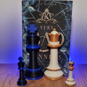 Könnte beinhalten: Schachspiel mit einem schwarzen und blauen König, einer weißen und goldenen Königin und einem kleinen schwarzen und blauen König und einer kleinen weißen und goldenen Königin. Die Schachfiguren stehen vor einem Buch mit dem Titel "VERY BAD ANNI".