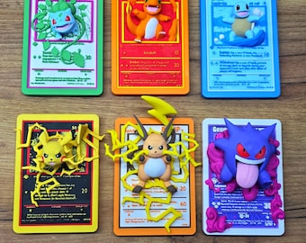 May include: Six colorful Pokémon trading card replicas featuring Bulbasaur, Charmander, Squirtle, Pikachu, Raichu, and Gengar. Each card has a 3D figure of the Pokémon and is set against a matching colored background. The cards are arranged on a wooden surface.