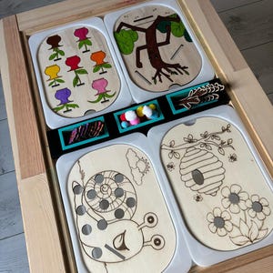 Peut inclure: Quatre planches d'activités en bois avec des thèmes différents : un jardin de fleurs, un arbre avec des racines, un escargot et une ruche. Chaque planche a des trous pour insérer des chevilles ou d'autres petits objets. Les planches sont posées sur une table en bois avec un plateau noir et un plateau turquoise contenant des chevilles colorées.