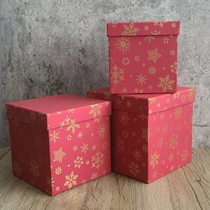 May include: Three square, lidded gift boxes in varying sizes. The boxes are a vibrant pink with a gold snowflake and star pattern. The boxes are stacked on a wooden surface against a textured gray wall.