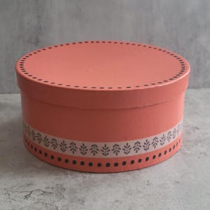 May include: A round, coral-colored storage box with a lid. The lid features a black dotted border along the edge. The side of the box is decorated with a horizontal band containing a repeating gray leaf pattern, framed by two rows of black dots.
