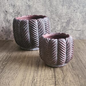 May include: Two decorative planters with a textured, woven design. The planters are a muted reddish-brown color with a lighter, weathered finish. The interior of the planters is a soft pink. They sit on a wooden surface.