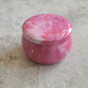 May include: A small, round, pink candle tin with a matching lid. The lid and body feature a marbled design in shades of pink and white. The tin is a solid pink color with small white speckles.