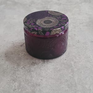 May include: A round, dark purple candle jar with a decorative floral pattern embossed on the glass. The lid is black with a gold and purple floral design. The jar sits on a light gray surface.