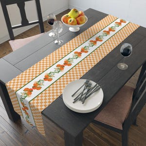 May include: A table runner with a white center stripe featuring orange fruit and floral designs, bordered by a green line. The runner is set on a dark wooden table with a checkered orange and white pattern. The table is set with plates, forks, and wine glasses.