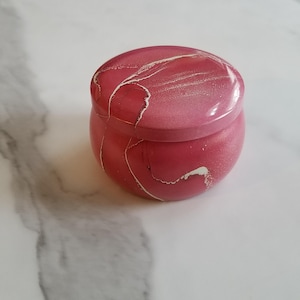 May include: A small, round, pink jar with a lid. The jar has a marbled effect with white streaks. The jar is sitting on a white and gray marble surface. The jar is likely used for storage or as a decorative item.