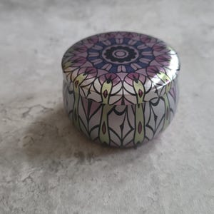 May include: A round, silver-colored tin with a decorative lid. The lid features a symmetrical floral design in shades of purple, pink, and blue. The sides of the tin have a matching pattern in silver and light green.