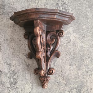 May include: A dark brown, ornate wooden wall shelf with a hexagonal top and decorative scrollwork. The shelf is designed to be mounted on a wall and has a detailed, carved appearance, showcasing a vintage aesthetic.