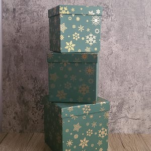 May include: Three stacked square gift boxes in varying sizes. The boxes are a deep teal green with a repeating pattern of gold snowflakes and stars. The boxes are arranged in a vertical tower.