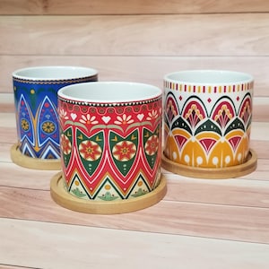 May include: Three ceramic plant pots with colorful geometric patterns. The pots sit on round wooden coasters. One pot is blue with yellow and white designs, another is red with green and yellow designs, and the third is white with yellow, red, and black designs.
