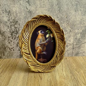 May include: Oval gold-colored picture frame with a detailed feather design. The frame surrounds a small painting of a monkey holding flowers. The monkey is orange and brown with a black face. The background is dark.