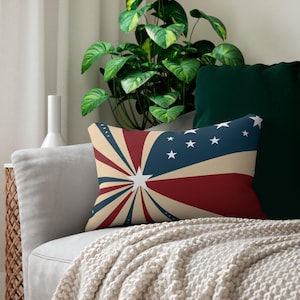 May include: Rectangular cushion with a patriotic design. The cushion features a starburst pattern in red, white, and blue, with white stars on a blue field. The cushion is placed on a grey sofa with a cream-coloured blanket.