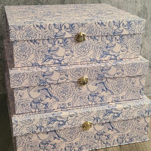 May include: A stack of three decorative storage boxes. Each box is covered in a light pink and blue floral and bird pattern. The boxes have gold-colored clasps. The boxes are nested, with the largest box at the bottom.