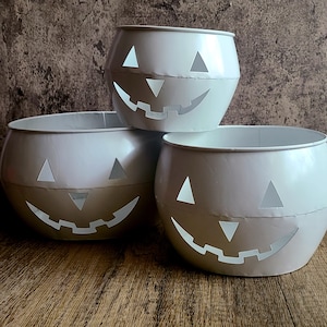 May include: Three white metal Halloween pumpkin-shaped containers stacked on a wooden surface. Each container has triangular eye cutouts and a jagged smile cutout. The containers are a light, matte white color.