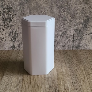 May include: A white, hexagonal container with a matching lid. The container is tall and slender, with a clean, minimalist design. It sits on a wooden surface against a textured gray wall.