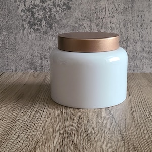 May include: A white ceramic jar with a rose gold lid. The jar has a rounded shape and a glossy finish. The lid is flat and circular. The jar is sitting on a wooden surface against a gray textured background.